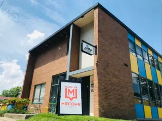 Modern brick building with large windows and a sign that reads "Made in Midtown" outside, under a blue sky with clouds.