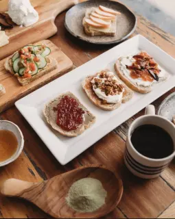 Assorted open-faced sandwiches with various toppings on a white platter, a small bowl of honey, a green powder in a wooden spoon, a cup of black coffee, and additional bread and toppings on a wooden t