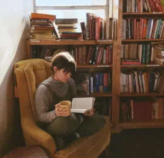 A person sitting on a yellow vintage armchair in a cozy room, reading a book and holding a yellow mug, with a bookshelf filled with books behind them and a window above the bookshelf letting in natura