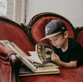 Young boy lying on a red velvet vintage sofa, reading a comic book titled 'Diary of a Minecraft Zombie', wearing a black shirt and a New Orleans Saints baseball cap.