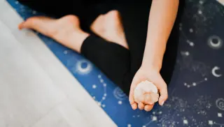 A person is sitting on a blue yoga mat adorned with celestial patterns, holding a crystal in their open hand, with bare feet visible in a relaxed pose.