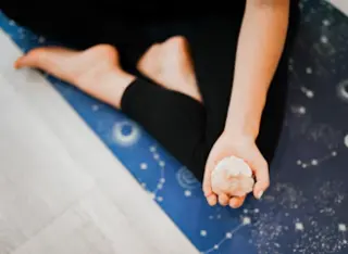 A person is sitting on a blue yoga mat adorned with celestial patterns, holding a crystal in their open hand, with bare feet visible in a relaxed pose.