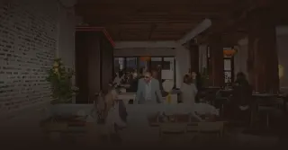 The dining room at Ila's Chicago: exposed brick, timber ceiling beams and warm pendant lights, with guests seated at tables and the bar behind them