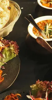 A spread of Ila's dishes on a dark table: flatbread in a wire basket, a curry in a copper serving pot, and plates finished with mixed greens
