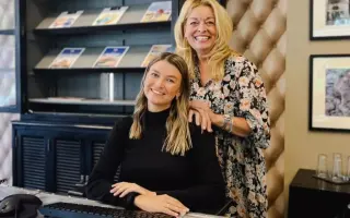 Zwei Reisebüromitarbeiterinnen im Büro, eine sitzt am Schreibtisch mit Tastatur, die andere steht dahinter vor einem Regal mit Reisekatalogen.