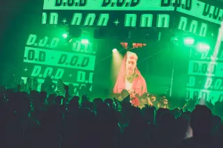 A DJ performs for an audience with green lighting and his name, D.O.D., displayed throughout the backdrop