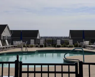 Outdoor swimming pool with lounge chairs and umbrellas, surrounded by a black fence, with house rooftops and a distant city skyline under a partly cloudy sky.