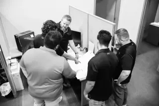 Six men gathered around a desk in an office, looking at documents and discussing projects for civil engineering, land entitlements, land surveying, construction quality assurance and quality control, 