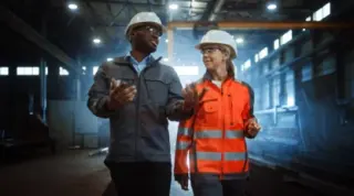A group of six men standing in a factory, engaged in conversation. One man in a white shirt holds a tablet. They are surrounded by industrial machinery and equipment.