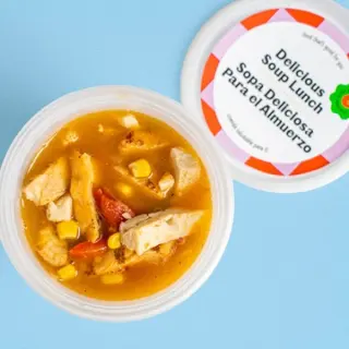 Container of chicken soup with vegetables and broth, next to a lid labeled 'Delicious Soup Lunch' in multiple languages.