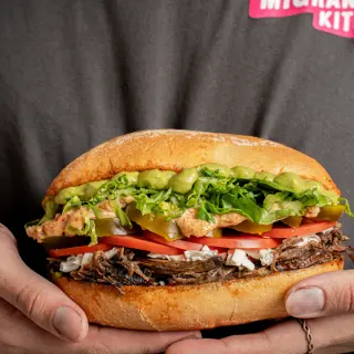 Close-up of a person holding a large sandwich with shredded beef, lettuce, tomato, pickles, avocado, and other ingredients in a hoagie bun.