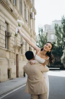 Jeune mariée en robe blanche et gants longs, soulevée par son époux dans une rue élégante, célébration d’un mariage raffiné organisé par Malia Agency