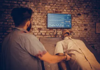 Een arts die een patiënt beoordeelt op een ziekenhuisbed met monitor die vitale functies weergeeft.
