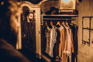 Een persoon kijkt in een oude houten kast gevuld met verschillende kledingstukken, waaronder jurken, jassen en shirts.