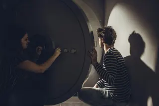 Een jonge vrouw en een jonge man kijken samen naar een wasmachine in een wasruimte, met een spil in de machine. Ze lijken te lachen en praten.