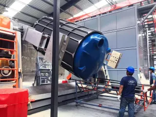 Workers in a factory maneuver a large industrial machine with a blue cylindrical component under a metal frame and overhead crane system.