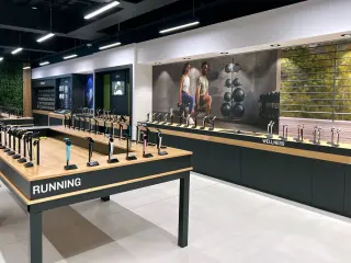 Display of fitness watches on wooden tables labeled 'RUNNING' and 'WELLNESS' inside a modern gym or sports store.