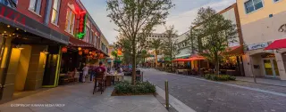 A lively outdoor shopping and dining street at sunset, with storefronts, tables, chairs, trees, and colorful lights. Clematis Street, Photography by Sunman, Sunman Media Group