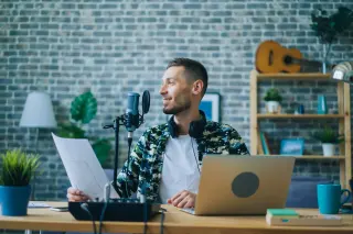 A man speaking into a microphone with headphones around his neck, sitting at a desk with a laptop, paper, and a coffee mug, in a room with a brick wall and shelves with books and decorative items. Pod