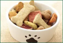 A white bowl with a black paw print contains assorted dog treats in various shapes, including bones and hearts.