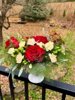 A floral arrangement with red roses, cream carnations, green hypericum berries, and greenery in a white vase on a black railing outdoors.