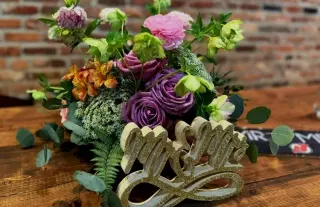 A colorful floral arrangement with various pink, purple, orange, and white flowers, green leaves, and a decorative gold 'Mrs. & Mr.' sign on a wooden table in front of a brick wall.