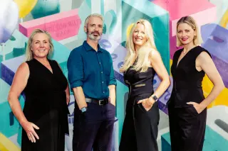 Four diverse adults, three women and one man, standing in front of a colorful graffiti wall, smiling at the camera.