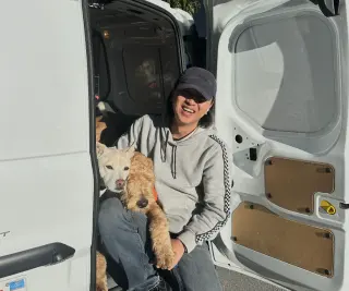 A person sitting inside the open door of a white van, smiling and holding two dogs, one white and one brown, with sunlight shining on them.