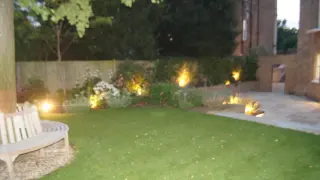 A blurry image of a backyard garden at dusk with a lawn, flower beds, and a paved pathway. There is a wooden bench on the left side and several outdoor lights illuminating the garden.