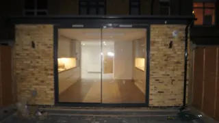 Front view of a small commercial or office space with a large glass sliding door, interior illuminated with warm lighting, and brick exterior wall surrounding the entrance.
