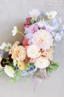 A colorful bouquet of various flowers, including dahlias, cosmos, and other blooms, arranged with green foliage and tied with a pale pink ribbon.