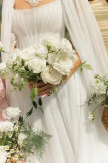 A bride in a white dress holding a bouquet of white roses, surrounded by other floral arrangements, outdoors.