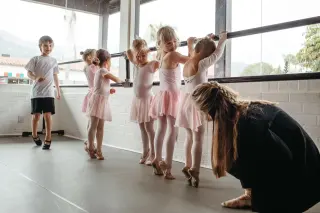 Ballet class with young girls wearing pink tutus and pointe shoes, with instructor helping on the floor, in a dance studio with large windows.