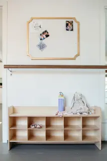 Empty wooden cubby shelf with a pair of pink shoes, a pink and white striped sock, a blue water bottle, a stuffed bunny, and a cloth on top, against a white wall with a bulletin board.