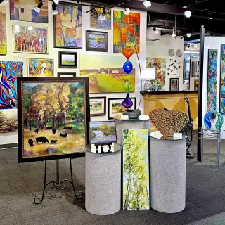 An art gallery display with various landscape and abstract paintings on white walls, with sculptures on a display tables. Overhead lighting highlights the artwork.