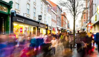 Busy city street with people walking and storefronts, including Superdrug and other shops, during daytime.