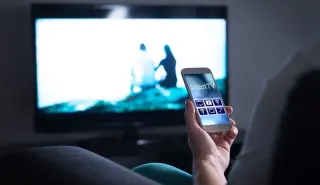 Person holding a smartphone with a smart TV remote app, sitting on a couch in front of a large flat-screen TV displaying a blurred scene of two people.